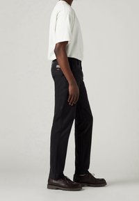 Uomo che indossa pantaloni neri a gamba dritta, una camicia bianca a maniche corte e scarpe stringate marrone scuro, in piedi davanti a uno sfondo semplice.