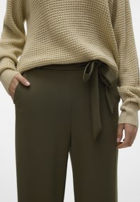 Pull en tricot beige avec un motif texturé, porté avec un pantalon vert olive doté d'une ceinture nouée et de poches latérales.