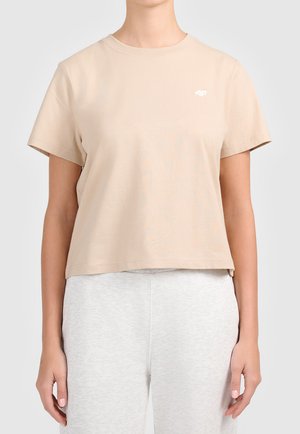 Personne portant un t-shirt beige à manches courtes avec un petit logo blanc sur la poitrine et un pantalon gris clair sur un fond uni.