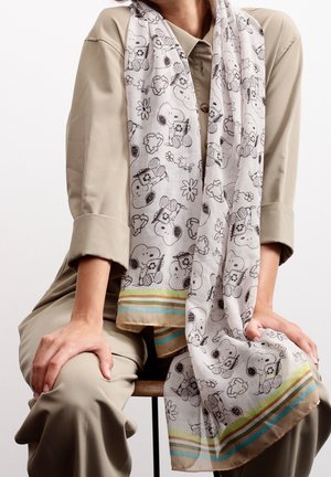 Écharpe légère avec croquis noir et blanc de Snoopy et fleurs, bord rayé vert, bleu et marron portée sur une chemise beige et un pantalon.