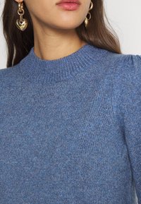 Maglione blu lavorato a maglia con collo alto a coste, caratterizzato da spalle a sbuffo. Sono visibili orecchini a forma di cuore dorati.