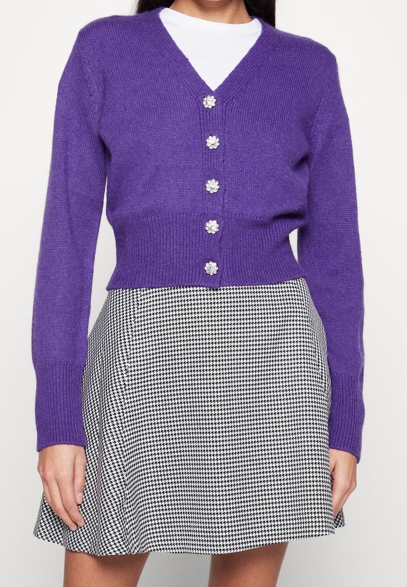 Lila gestrickte Strickjacke mit V-Ausschnitt, fünf blumenförmigen Knöpfen und langen Ärmeln, kombiniert mit einem schwarz-weißen Hahnentrittrock.