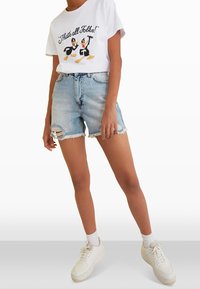 Camiseta gráfica blanca con dos personajes de dibujos animados, combinada con shorts de denim desgastados azul claros y zapatillas blancas. Conjunto casual de verano.