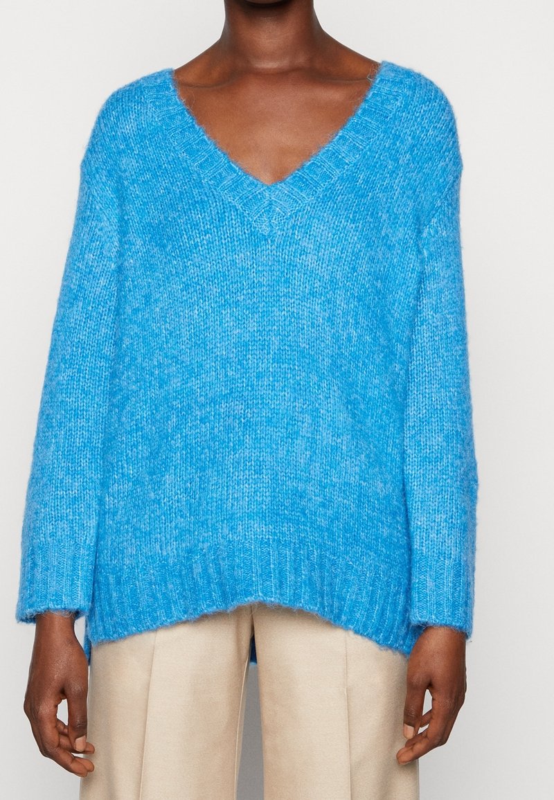 Persona di pelle scura che indossa un maglione V-neck ampio e peloso di un brillante blu e pantaloni beige, in piedi su uno sfondo bianco semplice.