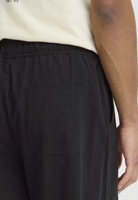 Sorte bukser i stof med elastisk talje, der har en enkelt laplomme bagpå. Materialet ser blødt ud med en glat tekstur.