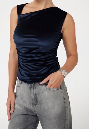 Woman wearing sleeveless navy blue velvet top and gray speckled high-waist jeans, with hand in pocket and silver wristwatch visible.