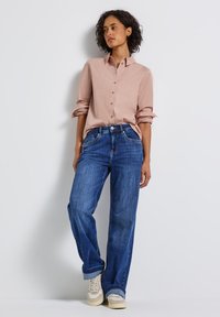 Femme aux cheveux bouclés portant une chemise rose poussiéreux boutonnée, un jean bleu ample retroussé aux chevilles, et des baskets beiges, debout contre un mur blanc.