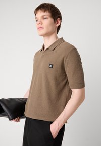 Brązowa polo z krótkim rękawem wykonana z teksturowanej tkaniny, z klasycznym kołnierzykiem i małą czarną naszywką z logo na piersi.