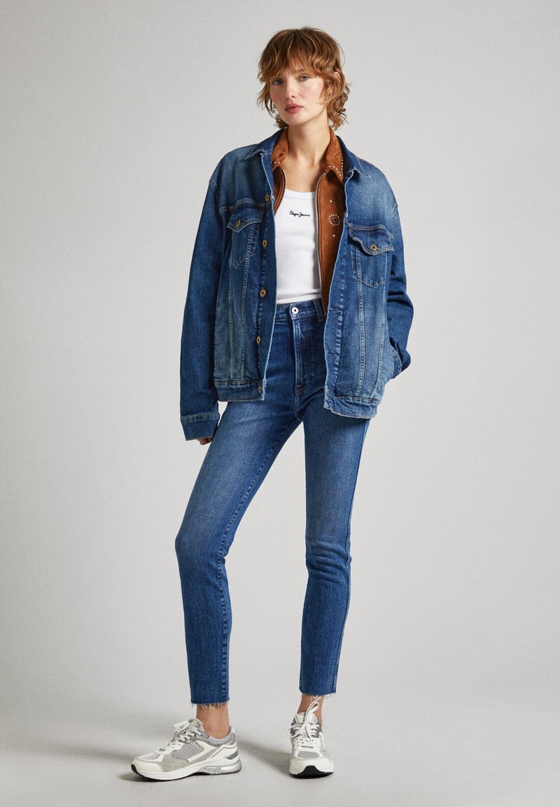 Chaqueta de denim y jeans de tiro alto en tonos azules variados, combinados con una camiseta sin mangas blanca. Las zapatillas presentan acentos en gris y blanco.