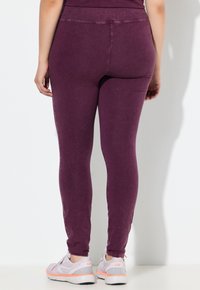 Leggings aderenti di un viola scuro, realizzati in tessuto morbido con una texture liscia. Presentano una vita alta e un design lungo fino alla caviglia. Abbinati a sneakers chiare.