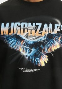 Melnas kokvilnas T-krekls ar lielu ērļas grafiku, kas ir ar ziliem spalviem, apkārt kam ir lodītes un zibens efekti, ar baltu tekstu.