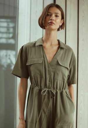 Kvinna med kort brunt hår som bär en grön byxdress med knappar, framfickor och midjeband, stående inomhus mot en trävägg.