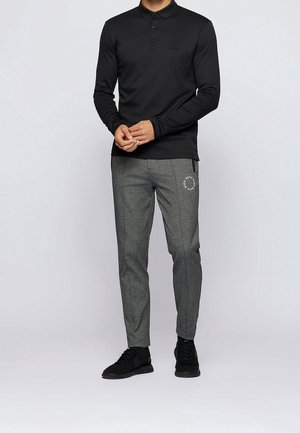 Polo noir à manches longues avec col boutonné, associé à un pantalon gris à motifs avec un accent logo, porté avec des baskets noires.