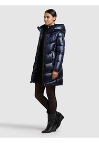 Tamsiai mėlynas pūkinis paltas su gaubtu, blizgiu paviršiumi ir užtrauktuku priekyje. Ilgos rankovės, šoninės kišenės, ilgio iki kelių kirpimas. Juodi aukštakulniai batai.