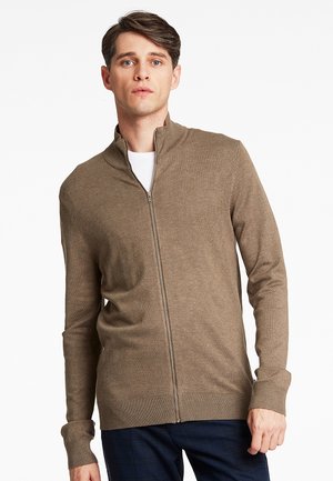Bruine ritstrui van zachte stof, met een hoge kraag, ribbelmanchetten en een getailleerde pasvorm, gedragen over een wit shirt.