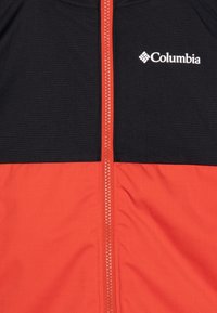 Columbia jakna s crnim gornjim dijelom, svijetlo crvenim donjim dijelom, središnjim patentnim zatvaračem i logotipom na lijevoj strani prsnog područja. Teksturirana tkanina.
