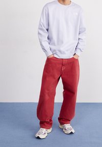 Osoba v světle fialové mikině, volných červených kalhotách a bílých teniskách stojící na modré podlaze proti jednoduché bílé zdi.