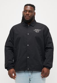 Crna lagana jakna s drukerima, šiljastim ovratnikom i izvezenim logotipom na prsima; ima elastične manžete i bočne džepove.