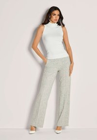 Vrouw in een wit mouwloos ribbed shirt, lichtgrijze gestreepte wijde broek, witte hakken, met één hand in de zak tegen een effen achtergrond.