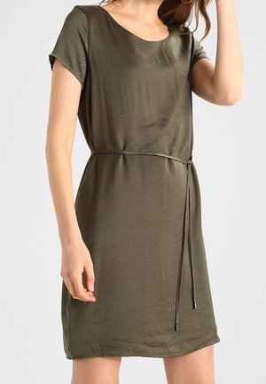 Kobieta ubrana w oliwkowozieloną sukienkę z krótkim rękawem i cienkim paskiem w talii, stojąca na tle jednolitego jasnego tła.