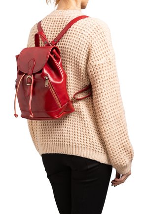 Sac à dos en cuir rouge avec un cordon de serrage, un rabat avant et deux zips sur les côtés. Porté par-dessus un pull en tricot beige.