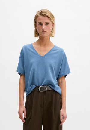 Femme blonde portant un haut bleu ample à col en V, rentré dans un pantalon marron foncé taille haute avec une ceinture noire, debout devant un fond blanc uni.