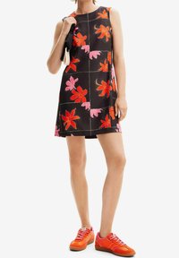 Černé bezrukávové šaty s jasnými červenými a růžovými květinovými vzory. Nosené s oranžovými teniskami, které ukazují kontrast v barvě a stylu.
