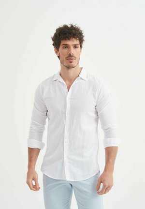 Chemise en lin blanc à boutons avec des manches longues retroussées aux poignets. Elle présente un col classique et une finition douce et texturée.