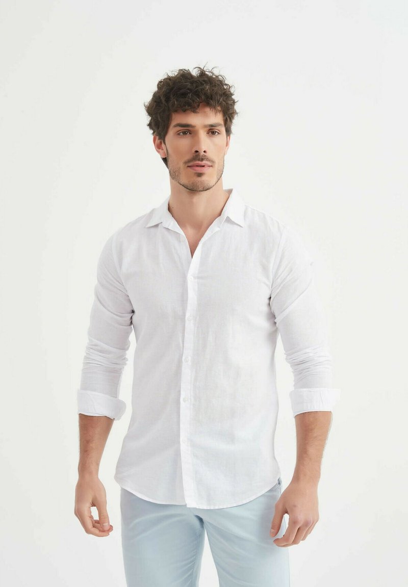 Chemise en lin blanc à boutons avec des manches longues retroussées aux poignets. Elle présente un col classique et une finition douce et texturée.