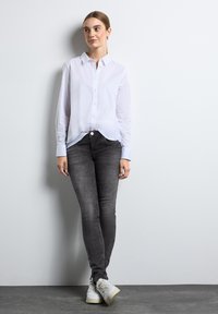 Vit skjorta med knappar, i kombination med slim fit grå jeans och vita sneakers. Modellen står mot en ljus bakgrund.