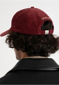 Burgundi värvi sametise pinnaga pesapalli müts reguleeritava rihmaga, brodeeritud logo ja sile metallic aktsent. Tekstuur on selgelt nähtav.