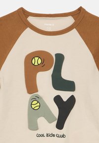 Μπεζ μπλουζάκι με καφέ raglan μανίκια, με πολύχρωμα γράμματα "PLAY" και τένις μπάλες, καθώς και το κείμενο "cool kids club" στο κάτω μέρος.