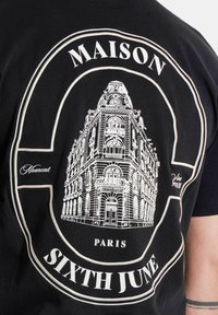 Zwarte t-shirt met een witte graphic van een gebouw, met de tekst "MAISON," "PARIS," en "SIXTH JUNE" in opvallende lettertypen.