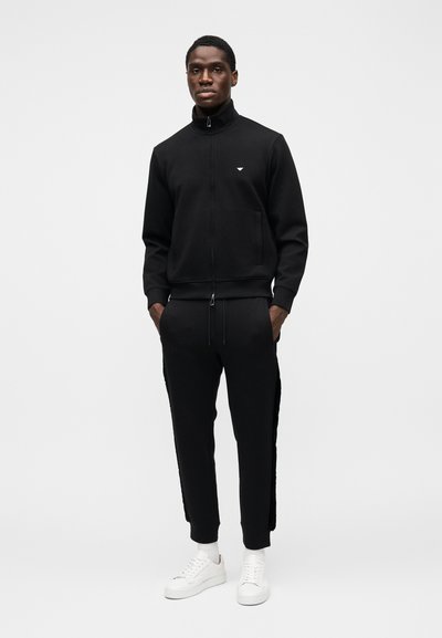 Svart treningsjakke med glidelås og matchende bukser med sidestreker, kombinert med hvite joggesko. Glatt stoff, tettsittende design, minimal merking.