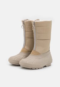 Botas de invierno beige con suelas de goma plana, parte superior de tela acolchada y cremalleras frontales. Presentan detalles de ante y textura acanalada en las suelas.