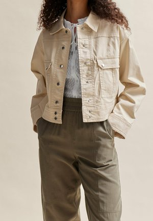 Kvinde iført en beige cropped jakke med trykknapper, en hvid bluse og olivengrønne højtaljede bukser med lommer.