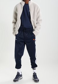 Chaqueta de forro polar en gris claro, pantalones deportivos azul marino con rayas laterales blancas, combinados con zapatillas grises y calcetines blancos. Diseño simple y deportivo.