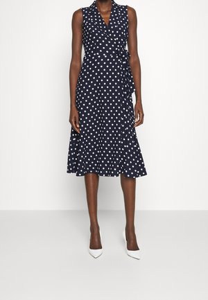 Robe portefeuille midi sans manches bleu marine à pois blancs, portée avec des escarpins blancs à bout pointu sur un fond blanc uni.