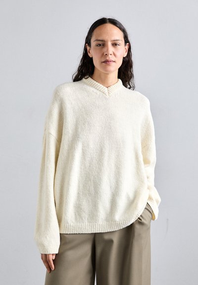 Cream, oversized knit sweater with a V-neck and ribbed detailing. Textured fabric and dropped shoulders, paired with beige trousers.