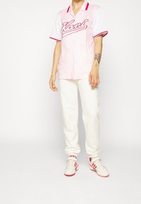 Różowo-biała koszula z krótkim rękawem o paskowanej fakturze i czerwonymi akcentami, zestawiona z białymi spodniami dresowymi i białymi tenisówkami z czerwonymi detalami.