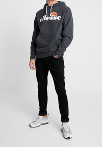 Felpa grigia con logo "ellesse" bianco e accento rosso. Presenta una tasca frontale e un cordino regolabile. Jeans neri slim-fit e sneaker bianche.