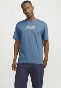 Blå bomulds T-shirt med korte ærmer og rund udskæring. Har et hvidt grafisk logo på brystet. Båret med mørkeblå denim jeans.