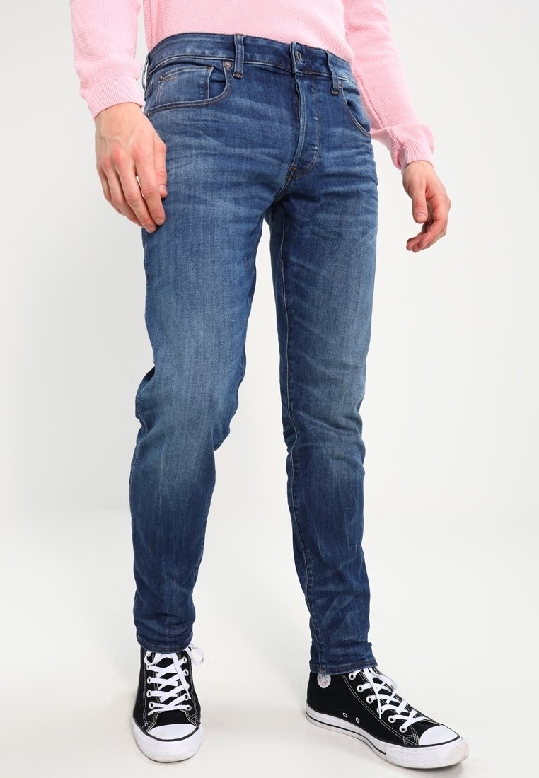 Eng anliegende blaue Denim-Jeans mit mittlerem Auswasch, Fünf-Taschen-Design und subtilen Ausbleichungen. Kombiniert mit schwarzen High-Top-Sneakern.