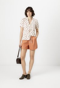Kortermet hvit bluse med rødt blomstermønster, bundet foran, parret med oransje shorts og grønne hæler, holder en brun veske.