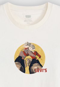 Bílý bavlněný tričko s kruhovou grafikou dvou cowboyů v károvaných a červených oblecích, se zápřahy a textem "LEVI'S" v červené barvě pod nimi.