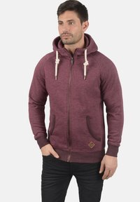 Sweat à capuche bordeaux zippé avec cordon de serrage, poches avant et poignets côtelés. Présente un petit patch logo marron sur l'ourlet inférieur.