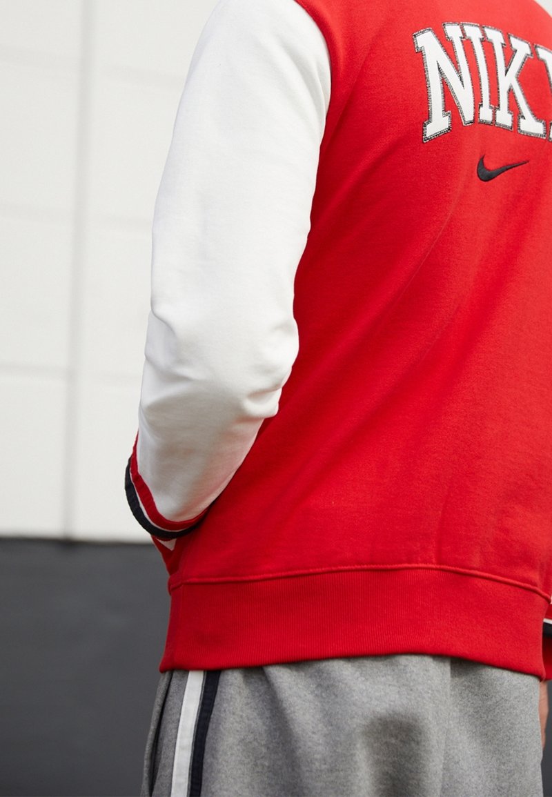 Sudadera roja con mangas blancas, con texto "NIKE" bordado. Textura suave, dobladillo acanalado y pantalones grises con rayas laterales.