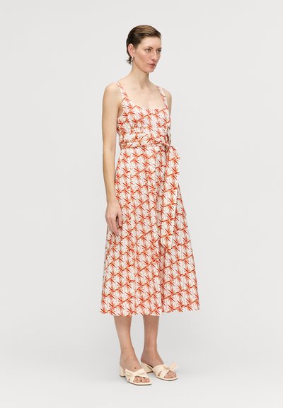 Maxi-Kleid aus weißem Stoff mit rotem Blättermuster, quadratischem Ausschnitt, verstellbaren Trägern und einer gebundenen Taillendetail. Getragen mit hellfarbigen Schuhen.