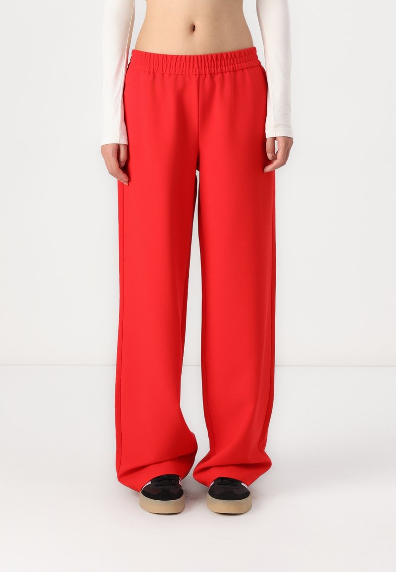 Personne portant un pantalon large rouge vif, des baskets noires et beiges, et un haut blanc à manches longues laissant apparaître le ventre sur un fond uni.