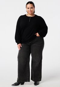 Vrouw staat in een zwarte langemouwen top, donkere wijde jeans, zwarte puntige schoenen, zilveren oorbellen en kettingen voor een witte achtergrond.